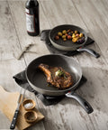 Two frying pans with cooked food on a wooden surface, including a bottle of wine and a cutting board.