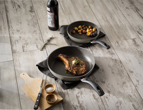 Two frying pans with cooked food on a wooden surface, including a bottle of wine and a cutting board.