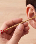 Person using a wooden ear cleaning tool with a red charm near their ear.