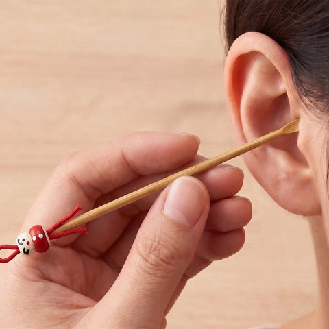 Person using a wooden ear cleaning tool with a red charm near their ear.