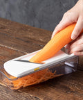 A hands-on demonstration of a white and clear mandoline slicer on a rustic wooden table, with a person slicing an orange carrot into shredded pieces.