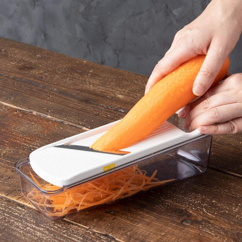 A hands-on demonstration of a white and clear mandoline slicer on a rustic wooden table, with a person slicing an orange carrot into shredded pieces.