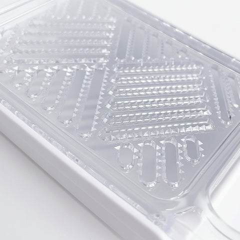 A close-up of a clear plastic grater with a white base, showing the textured grating surfaces and handle attachment, set against a white background.