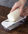 A hands-free mandoline slicer with a clear plastic container base, white blade cover, and stainless steel blade, shown with a white radish being pushed through by a person's hands on a dark wooden table.