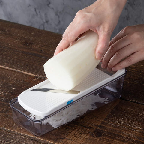 A hands-free mandoline slicer with a clear plastic container base, white blade cover, and stainless steel blade, shown with a white radish being pushed through by a person's hands on a dark wooden table.