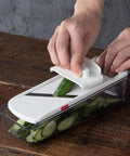 A hands-on demonstration of a kitchen mandoline slicer with a clear container, showing a cucumber being sliced on a dark wooden table.