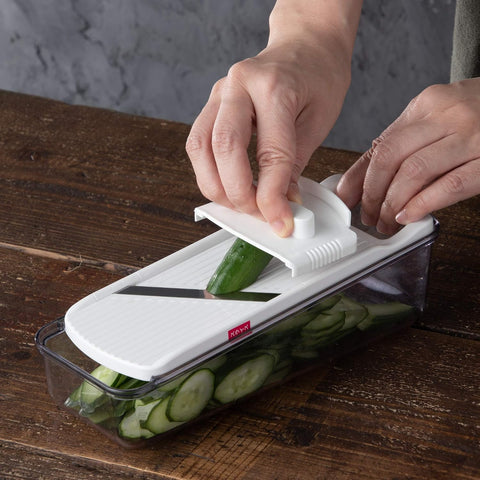 A hands-on demonstration of a kitchen mandoline slicer with a clear container, showing a cucumber being sliced on a dark wooden table.
