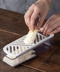 A hands-on demonstration of a white and clear plastic vegetable slicer on a rustic wooden table, with a person's hands guiding a piece of radish through the grater.