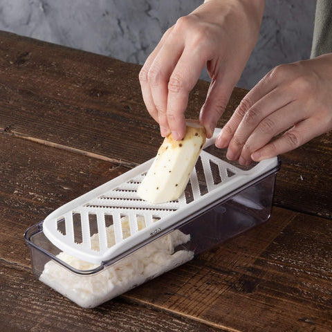 A hands-on demonstration of a white and clear plastic vegetable slicer on a rustic wooden table, with a person's hands guiding a piece of radish through the grater.