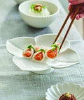 Four dumplings filled with orange-pink salmon and topped with green scallion, served on a white flower-shaped plate. A hand holding wooden chopsticks is lifting one dumpling.