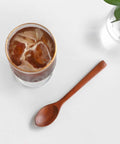 A clear glass tumbler with a gold rim filled with iced coffee and ice, placed on a white surface beside a wooden spoon and a small glass vase with green leaves.