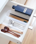 A white drawer organizer tray holding kitchen utensils, including a dark blue grater, chopsticks, forks, spoons, and wooden spoons, shown in a real drawer setting.