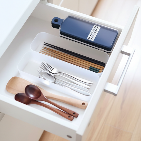 A white drawer organizer tray holding kitchen utensils, including a dark blue grater, chopsticks, forks, spoons, and wooden spoons, shown in a real drawer setting.