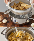 Pasta being drained in a stainless steel colander over a pot on a wooden surface.