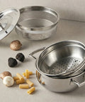 Stackable stainless steel steamer baskets and a pot on a neutral background with pasta and mushrooms