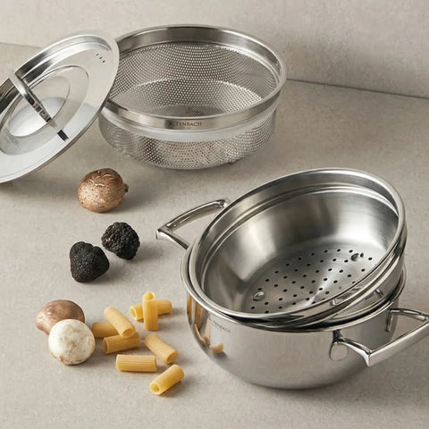 Stackable stainless steel steamer baskets and a pot on a neutral background with pasta and mushrooms