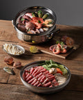 Two metal steamer baskets with raw seafood and vegetables on a wooden surface.
