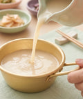 Person pouring Korean rice wine into a gold bowl on a wooden table.