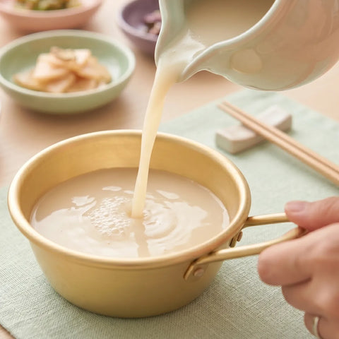 Person pouring Korean rice wine into a gold bowl on a wooden table.