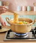 Person using chopsticks to lift noodles from a pot on a stove with a kitchen background.