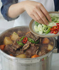 Stewing pot with beef and vegetables, person adding green onions and red peppers.