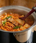 Stirring a pot of hot kimchi stew with a red spoon, featuring the cookware brand 'Altenbach'.