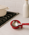 Red measuring cup with spoon on a kitchen counter next to a pot on a stove.