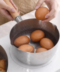 A stainless steel saucepan with a wooden handle is shown on a white electric cooktop, with several brown eggs being placed into the pot by hands. A second pot with food is partially visible on the left.