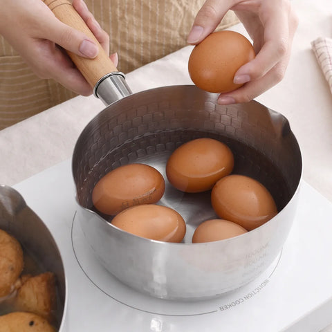A stainless steel saucepan with a wooden handle is shown on a white electric cooktop, with several brown eggs being placed into the pot by hands. A second pot with food is partially visible on the left.