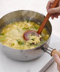 A stainless steel saucepan filled with a light yellow soup, green onions, and egg pieces, being stirred with a wooden-handled spoon on a white electric burner. A hand holds the spoon handle, and another hand holds the pan handle.