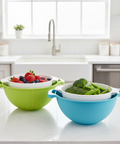 Two colanders, one green and one blue, on a kitchen counter with fruits and vegetables.