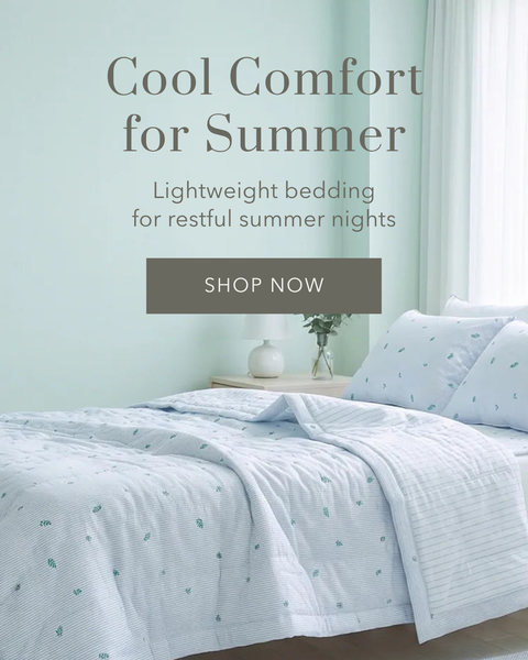 A bed dressed in a cool white bedding set with small scattered green leaf motifs, styled in a bedroom with a pale mint wall, a nightstand, and a window. Large text reads 'Cool Comfort for Summer' with a 'SHOP NOW' button.