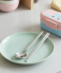 Light green plate with silver spoon and chopsticks, pink and blue bento box on a light surface.