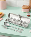 Stainless steel cutlery set in a case on a light green surface with food containers in the background.