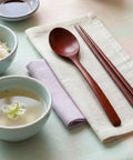 Wooden spoon and chopsticks on a folded cloth with a bowl of soup and small bowl of rice in the background.