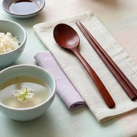 Wooden spoon and chopsticks on a folded cloth with a bowl of soup and small bowl of rice in the background.
