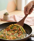Person using a wooden spatula to serve a Korean seafood pancake with vegetables from a frying pan on a table.