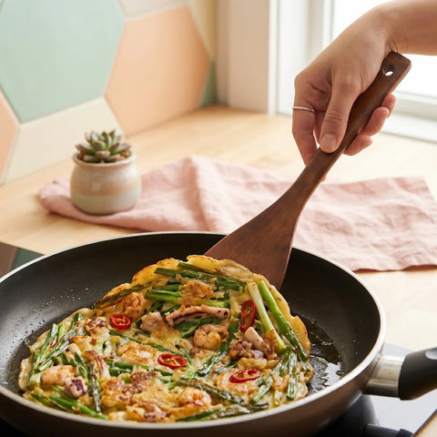 Person using a wooden spatula to serve a Korean seafood pancake with vegetables from a frying pan on a table.