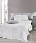 A neatly made bed in a modern bedroom setting, featuring white bedding with a delicate gray floral pattern on the comforter, styled with white and gray pillows against a gray headboard. A matching gray nightstand with a lamp is visible on the right.