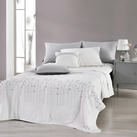 A neatly made bed in a modern bedroom setting, featuring white bedding with a delicate gray floral pattern on the comforter, styled with white and gray pillows against a gray headboard. A matching gray nightstand with a lamp is visible on the right.