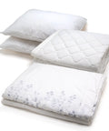 Three folded white bedding items stacked on a white background, including two pillows and a quilted mattress protector and comforter with a subtle floral pattern on the lower folded piece.