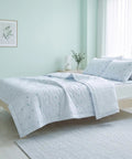 A light blue and white bedding set with a small leaf pattern, styled on a light wood bed frame in a bright, pale green bedroom.