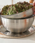 Stainless steel colander with lettuce being drained over a bowl on a kitchen counter.