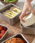 Person preparing a lunch with sandwiches and tomatoes in metal containers on a woven mat.