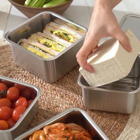 Person preparing a lunch with sandwiches and tomatoes in metal containers on a woven mat.