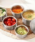 Set of stainless steel containers with various food items on a woven mat