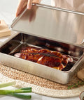Stainless steel roasting pan with roasted meat on a woven mat, surrounded by vegetables.