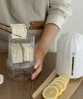 Person holding a container of tofu next to a white appliance on a wooden surface with lemons.