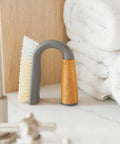 U-shaped cleaning brush with gray bristle head and wooden handle on a light surface with white towels in the background