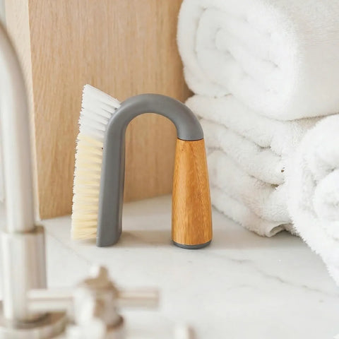 U-shaped cleaning brush with gray bristle head and wooden handle on a light surface with white towels in the background